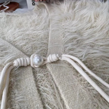 Close-up of a cream-colored knit garment with a knotted cord detail and a central faux pearl, laid flat on a fluffy cream faux-fur surface.