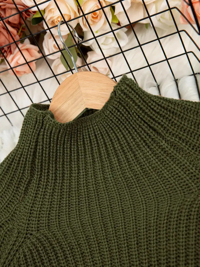 An olive green ribbed knit mock-neck sweater hanging on a light wood hanger, set against a white textured background with a black wire grid and peach roses.