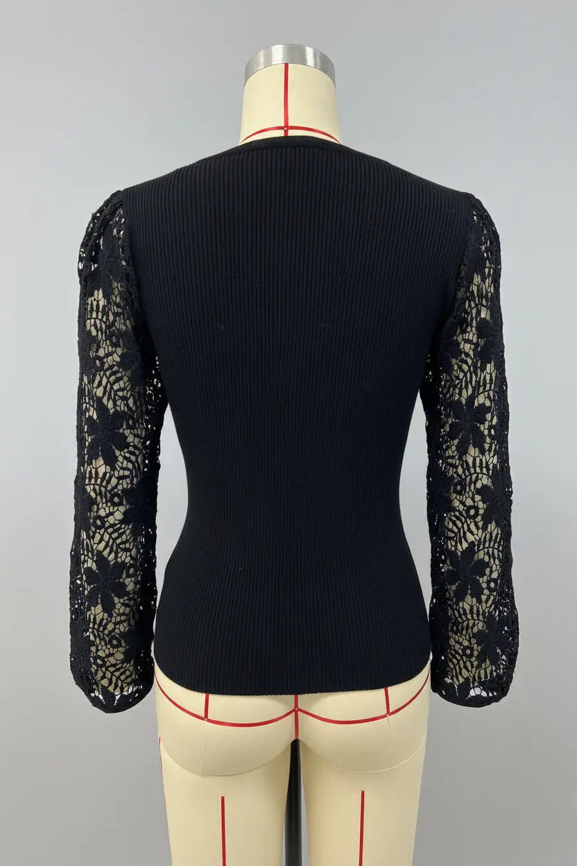 A black long-sleeve top with a ribbed body and sheer floral lace sleeves, displayed on a beige dress form against a white wall with framed city artwork.