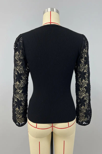 A black long-sleeve top with a ribbed body and sheer floral lace sleeves, displayed on a beige dress form against a white wall with framed city artwork.
