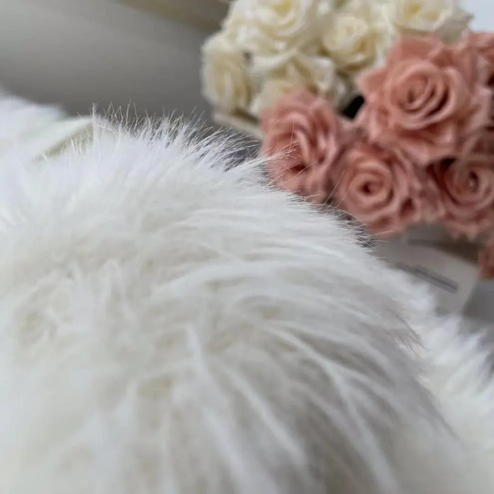 A close-up of a fluffy white faux fur or sheepskin material in the foreground, with soft pink and cream roses blurred in the background.