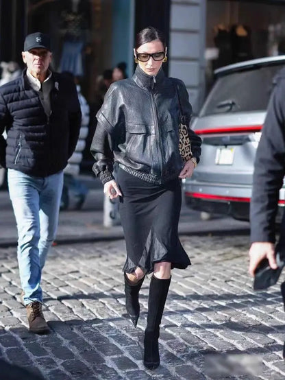 A woman walking on a cobblestone street wearing a black leather jacket, black skirt, black knee-high boots, and black sunglasses, with a leopard-print shoulder bag.