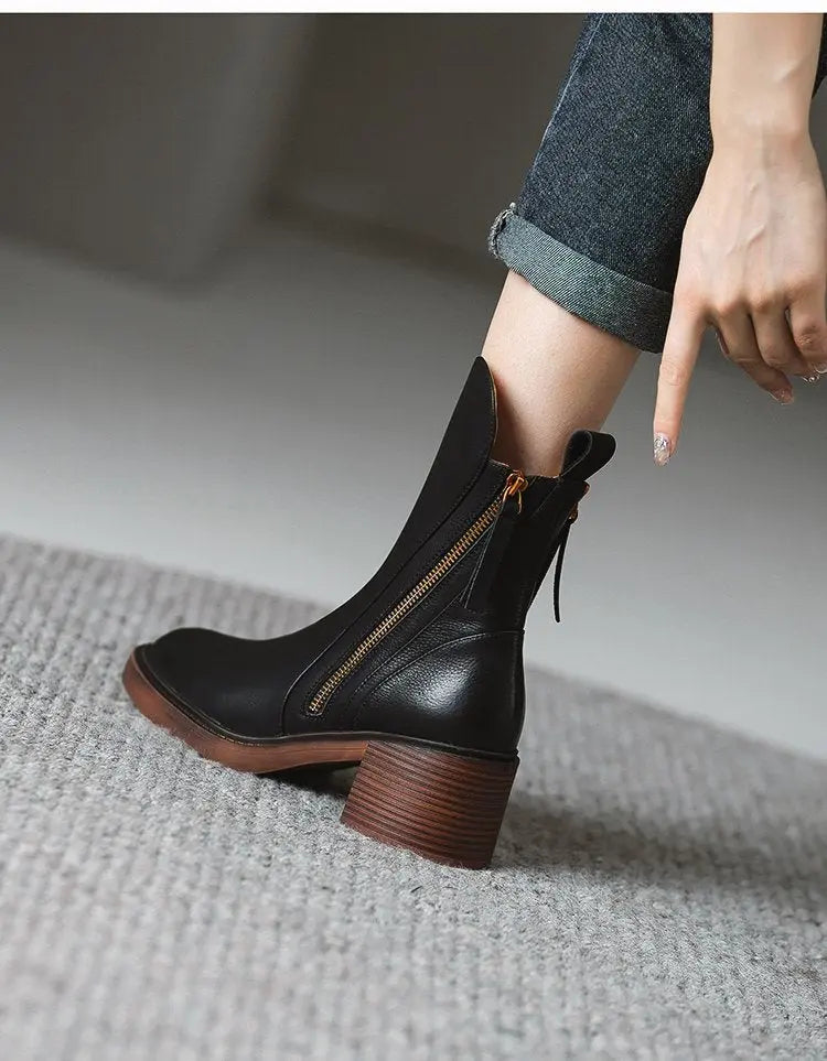 A person wearing a black leather ankle boot with a brown stacked block heel and a gold zipper, paired with cuffed dark gray jeans, pointing at the boot.
