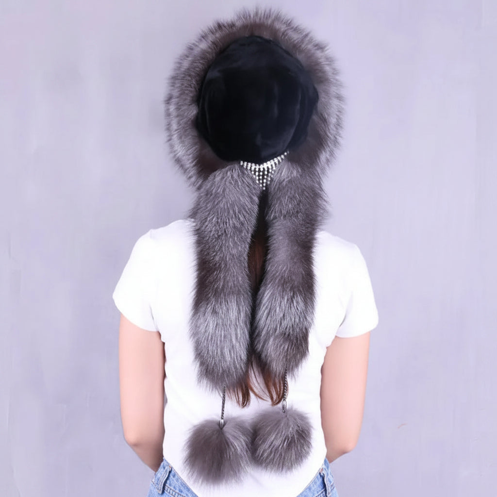 A woman shown from the back wearing a luxurious winter hat with a large gray and silver fox fur trim, a black felt crown, and two pom-poms at the bottom. The brand logo 'Jools cana' is visible in the top left corner.
