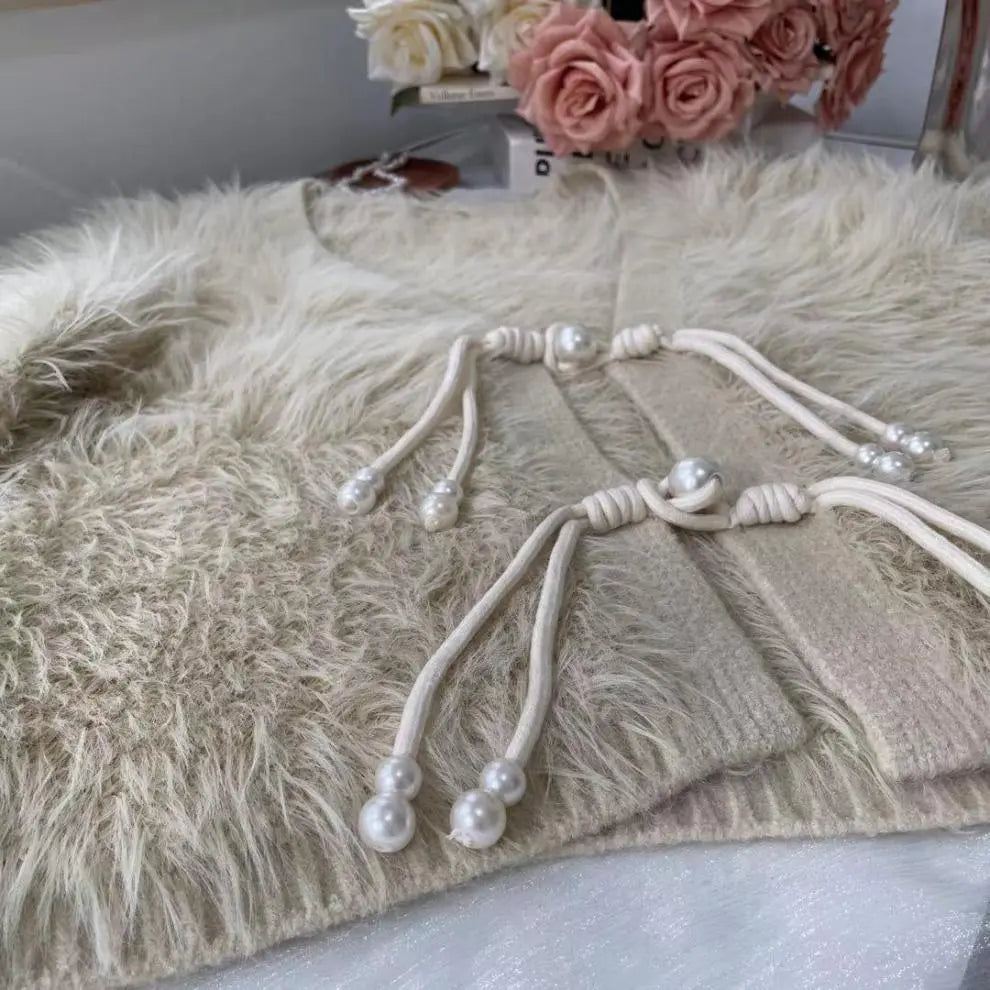 A beige faux fur cardigan with a cream-colored knotted cord and pearl bead details, styled on a white surface with pink roses in the background.