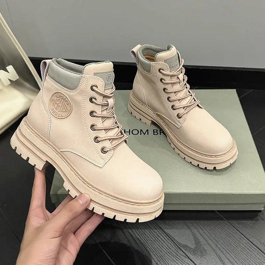 A pair of light beige lace-up ankle boots with chunky lug soles, shown with one boot being held by a hand and the other resting on a box. A shoebox is partially visible on the left.