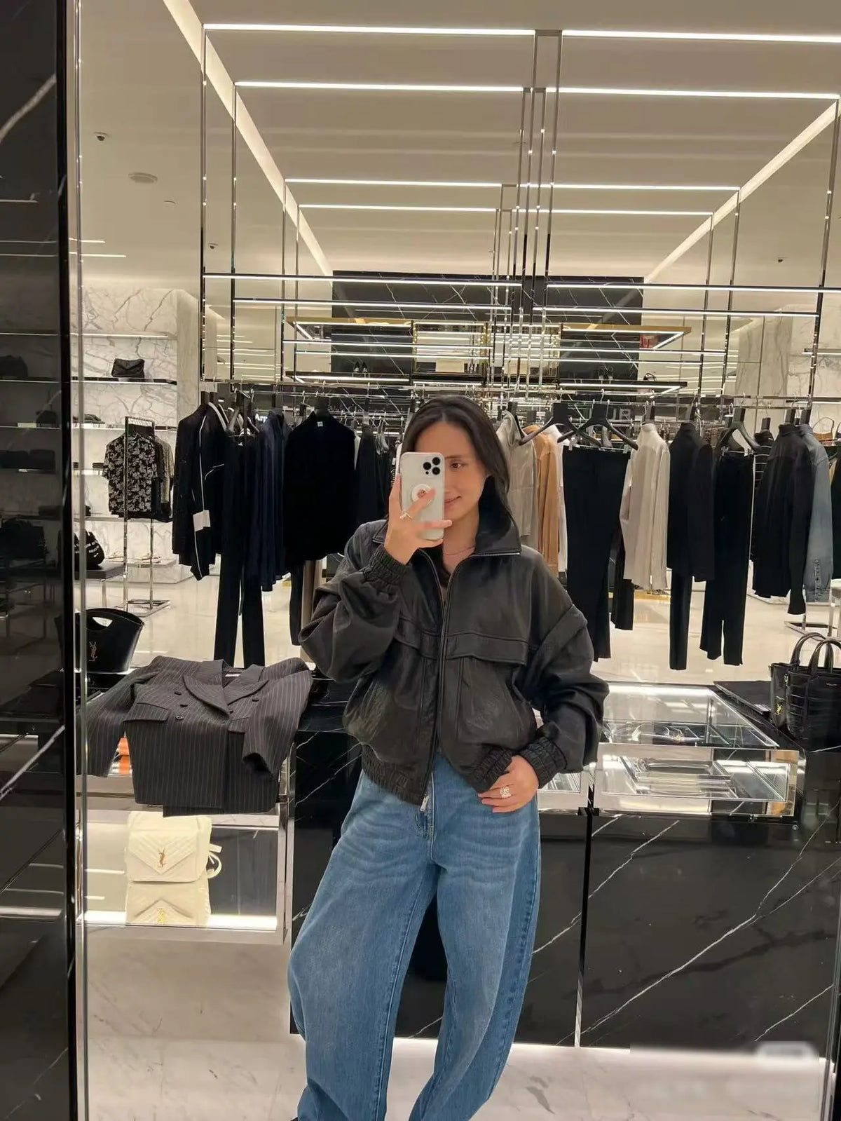 A woman takes a mirror selfie in a clothing boutique, wearing a dark gray oversized utility jacket and light blue wide-leg jeans.