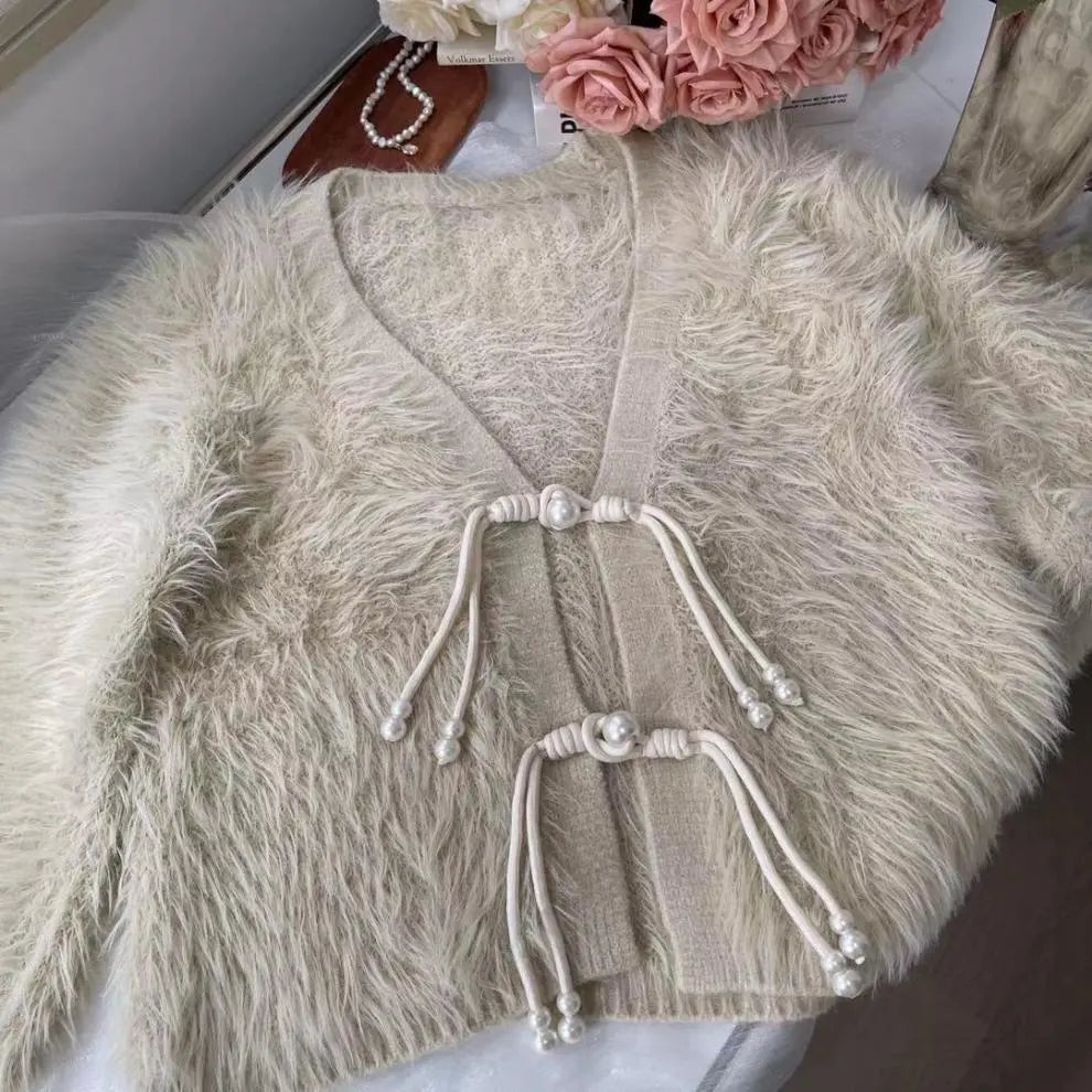 A cream-colored faux fur cardigan with a knit trim and decorative pearl-topped drawstring ties, laid flat on a light surface with pink roses in the background.