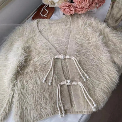 A cream-colored faux fur cardigan with a knit trim and decorative pearl-topped drawstring ties, laid flat on a light surface with pink roses in the background.