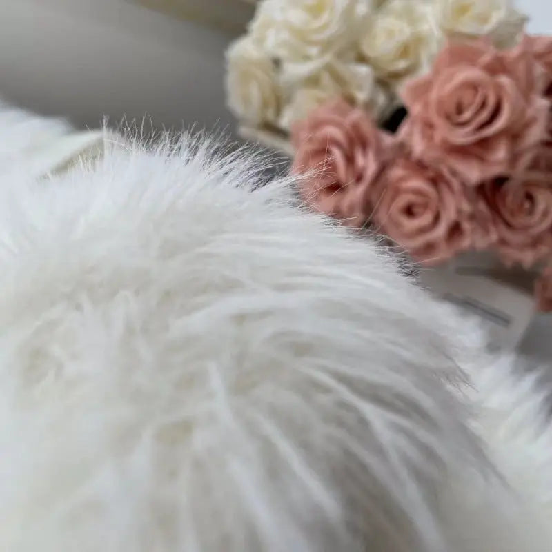 A close-up of a fluffy white faux fur or sheepskin-style textile in the foreground, with blurred cream and blush pink roses in the background.