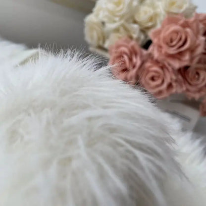 A close-up of a fluffy white faux fur or sheepskin-style textile in the foreground, with blurred cream and blush pink roses in the background.