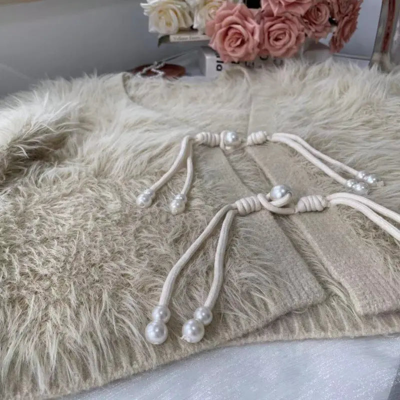 A beige faux fur cardigan with a cream-colored knotted cord and pearl bead details, styled on a white surface with pink roses in the background.