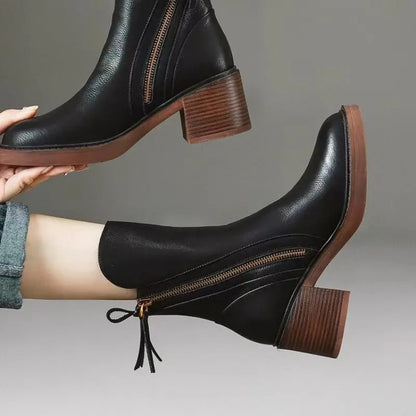 Black leather ankle boots with brown soles worn by a person on a gray background