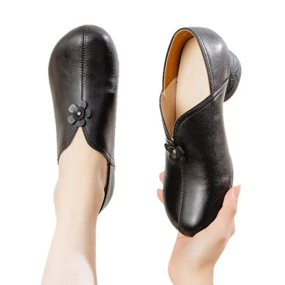 A pair of black leather slip-on shoes with decorative flower details, shown on a foot and held in a hand against a plain white background.