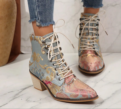 A pair of lace-up ankle boots with a light blue base and ornate gold, pink, and beige floral embroidery, shown on a marble floor with a person's jeans visible.