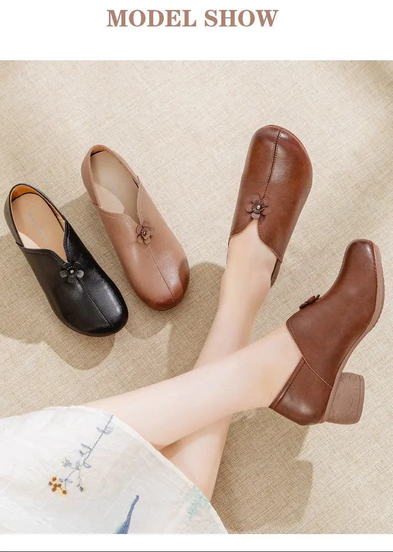 Three women's slip-on low-heel shoes displayed on a beige textured surface, with one shown on a model's foot. The shoes come in black, tan, and brown, each featuring a small decorative flower detail near the opening.