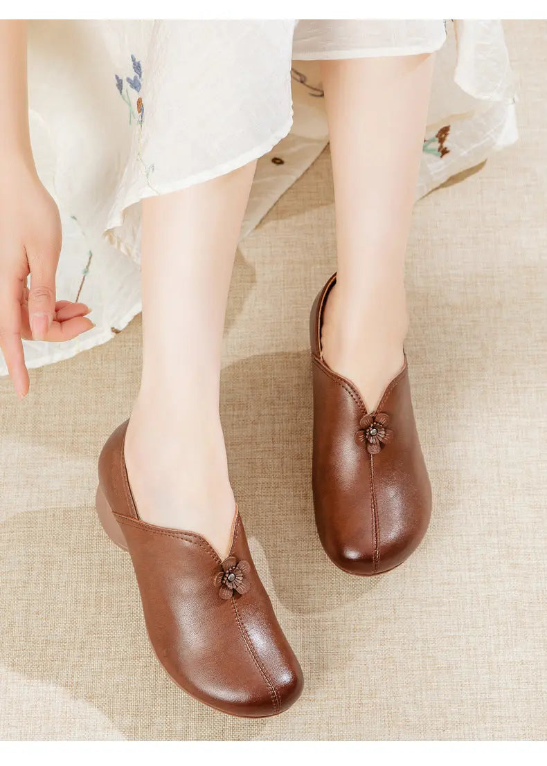 A person wearing brown leather slip-on shoes with decorative floral embellishments, seated on a light beige textured surface with a white floral skirt visible.