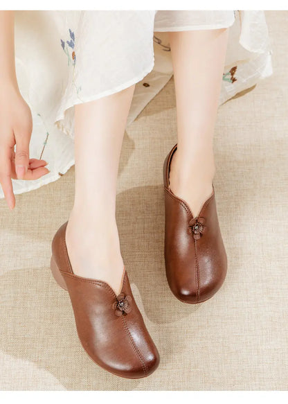 A person wearing brown leather slip-on shoes with decorative floral embellishments, seated on a light beige textured surface with a white floral skirt visible.