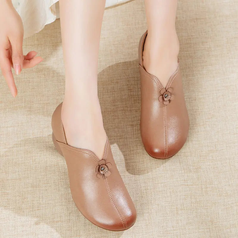 A person wearing a pair of brown slip-on mule-style shoes with small decorative floral embellishments near the front, standing on a beige textured surface.
