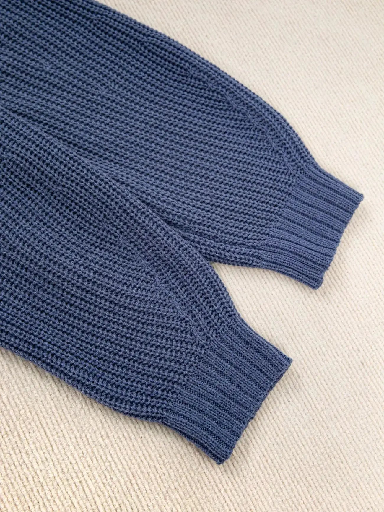 Close-up of a textured knit sweater sleeve with a ribbed cuff, laid flat on a light beige woven fabric background.