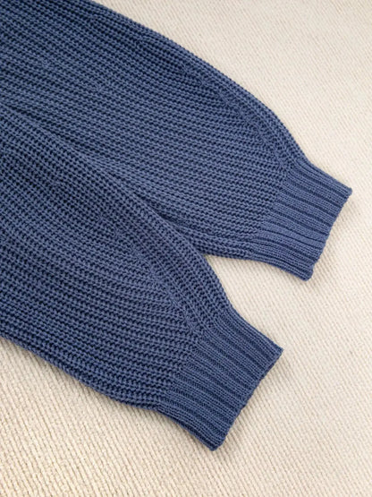 Close-up of a textured knit sweater sleeve with a ribbed cuff, laid flat on a light beige woven fabric background.
