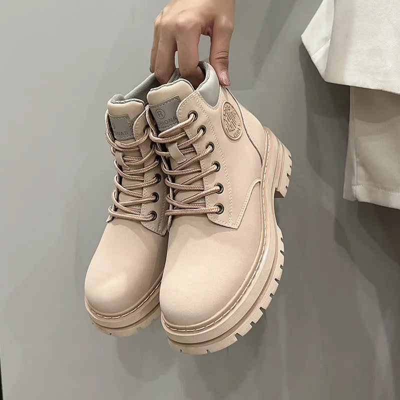 A pair of chunky platform ankle boots in a light beige color, held by a hand against a gray wall background.