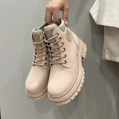 A pair of chunky platform ankle boots in a light beige color, held by a hand against a gray wall background.