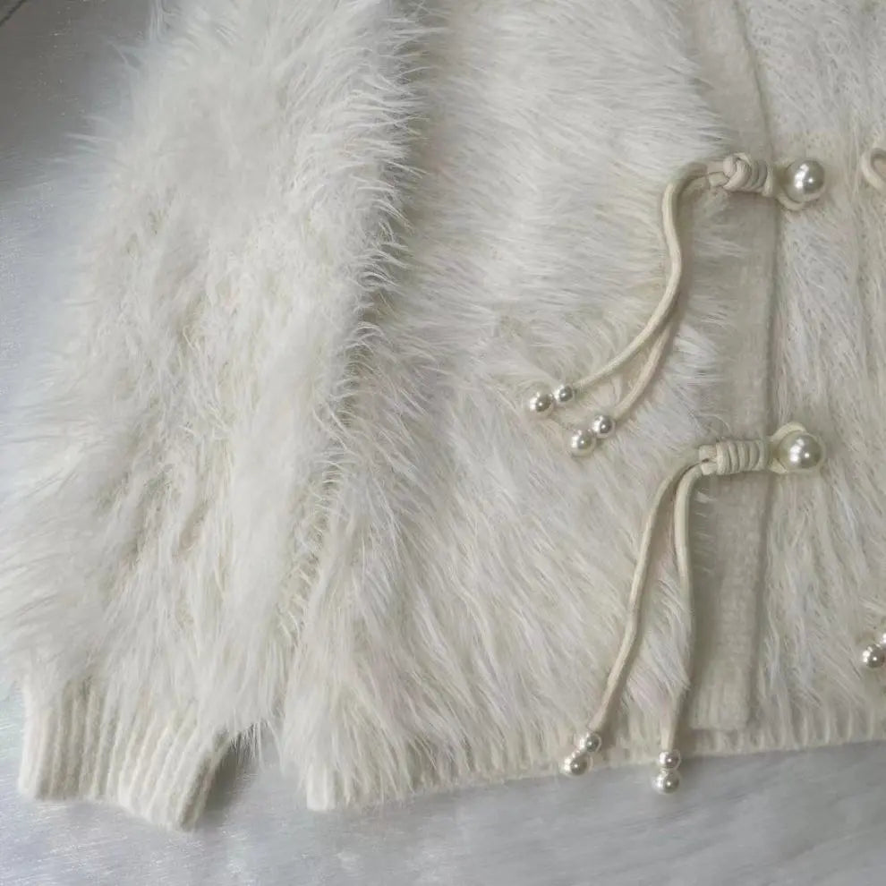 Close-up of a fluffy cream faux-fur cardigan with drawstring closures adorned with small pearl-like beads, laid flat on a light fabric surface.