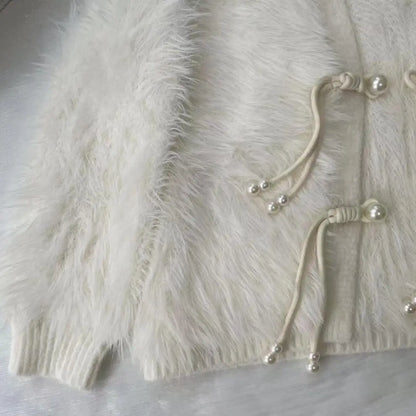 Close-up of a fluffy cream faux-fur cardigan with drawstring closures adorned with small pearl-like beads, laid flat on a light fabric surface.