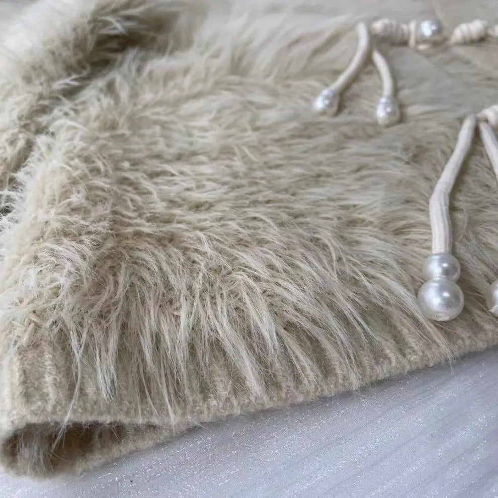 Close-up of a beige faux fur or sheepskin textile with long shaggy fibers and white cord drawstrings tipped with pearl-like beads, laid on a light surface.