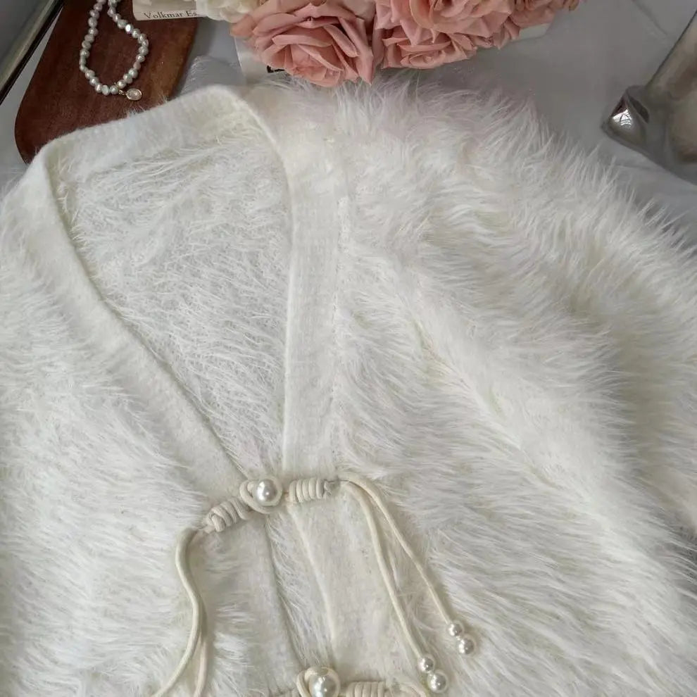 A close-up of a fluffy white faux fur garment, likely a cardigan or sweater, featuring a V-neckline and decorative cord ties with pearl-like beads. The garment is styled with a pearl necklace and pink flowers in the background.