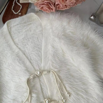 A close-up of a fluffy white faux fur garment, likely a cardigan or sweater, featuring a V-neckline and decorative cord ties with pearl-like beads. The garment is styled with a pearl necklace and pink flowers in the background.