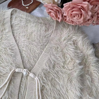 A close-up of a fuzzy knit cardigan in a warm off-white or cream tone, styled with pink roses and a pearl necklace on a white surface.