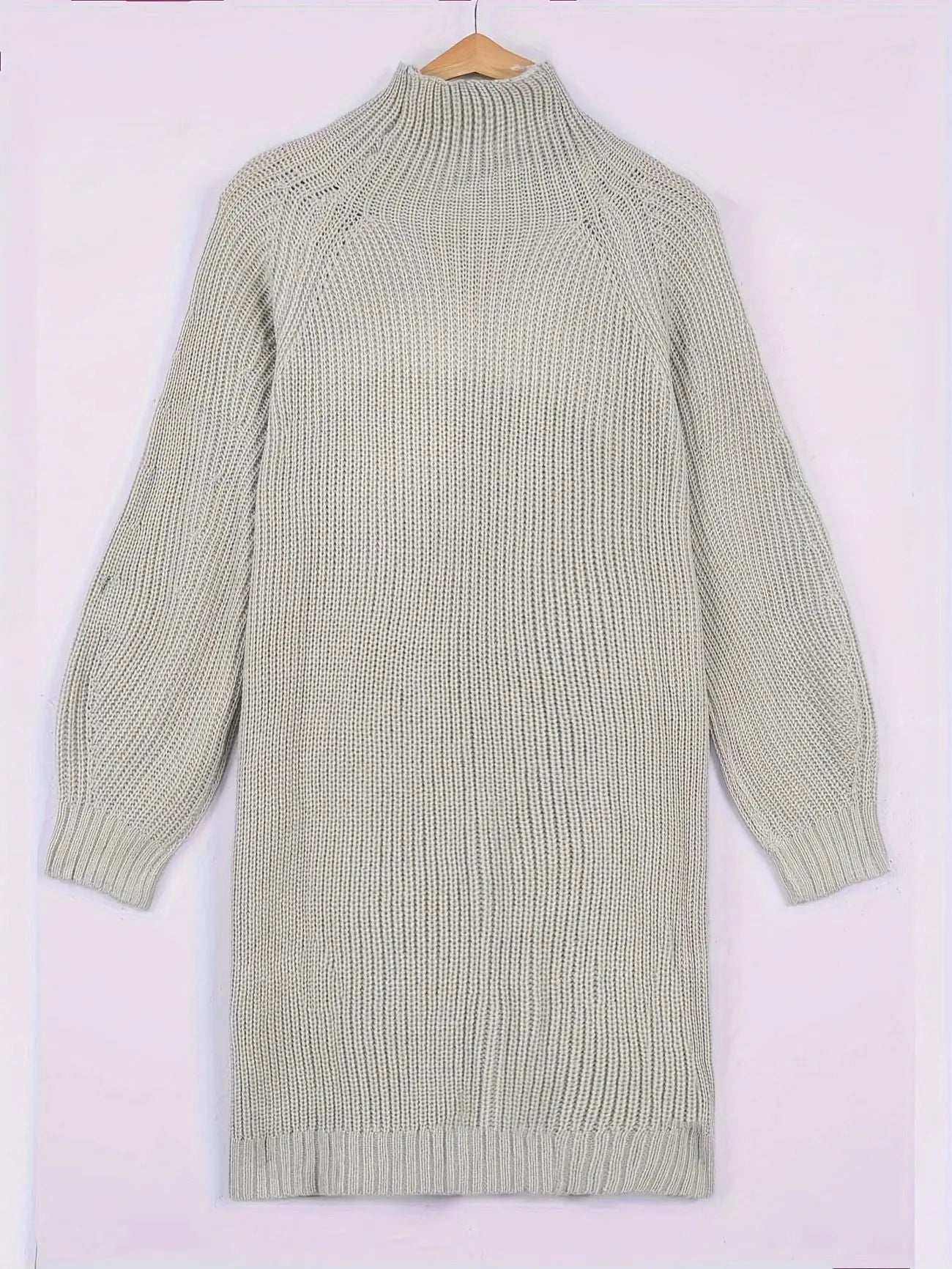 A light sage green ribbed knit mock-neck sweater dress with long raglan sleeves, hanging on a wooden hanger against a pale pink background.