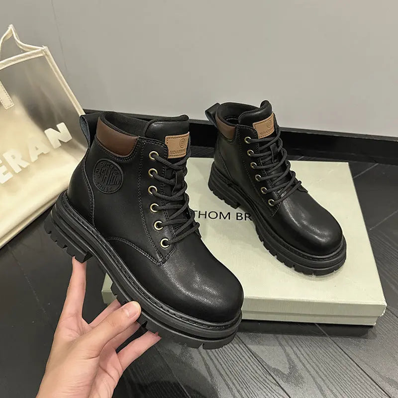 A pair of black leather lace-up ankle boots with brown heel tabs and gold-tone eyelets, shown on a dark floor with a shoe box and a hand holding one boot. The brand name THOM BROWN is partially visible on the box.