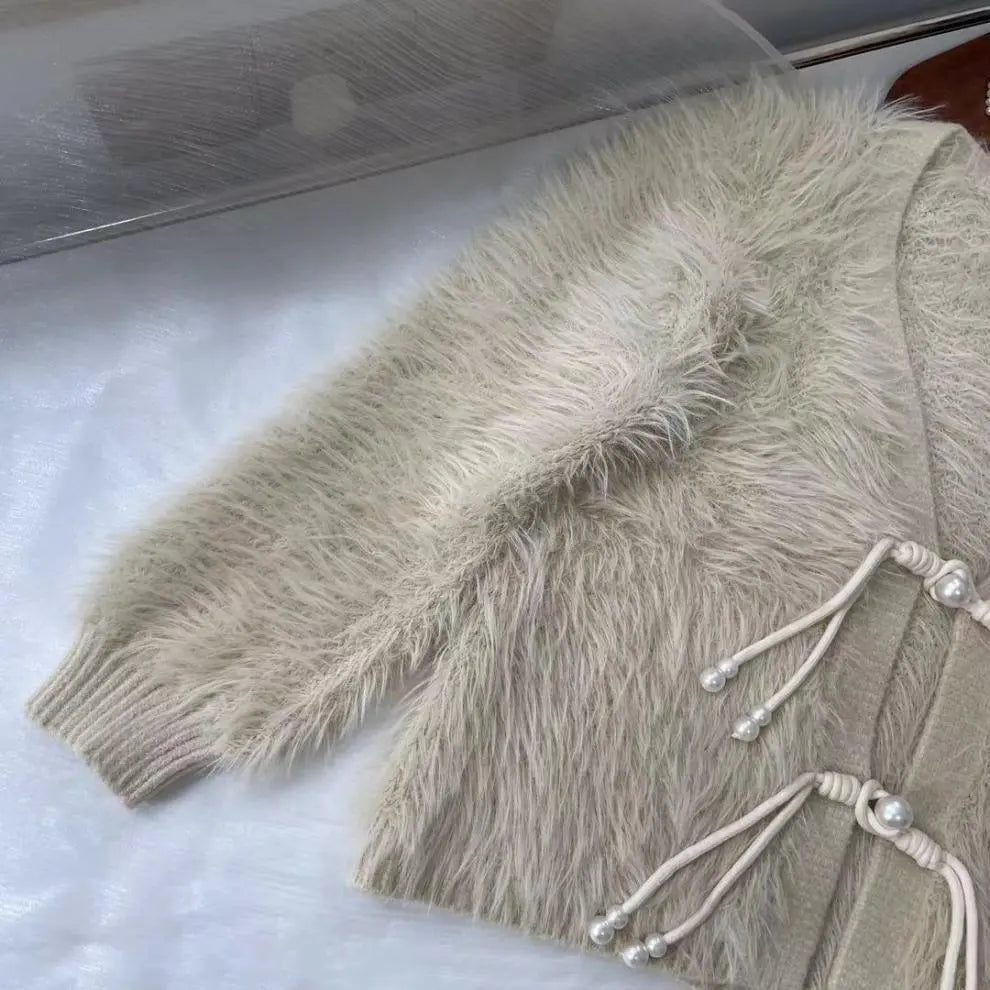A close-up of a beige faux fur cardigan with a ribbed knit collar, cuffs, and hem, featuring decorative cord and pearl details on the front, laid flat on a white surface.
