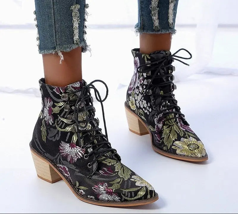 A pair of women's lace-up ankle boots with a black base and a colorful floral pattern, styled with frayed blue jeans. The boots feature a chunky light wood block heel and black laces.