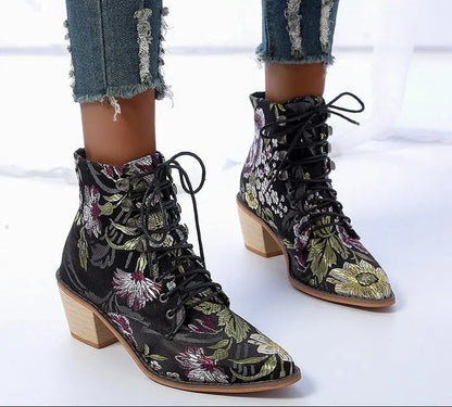 A pair of women's lace-up ankle boots with a black base and a colorful floral pattern, styled with frayed blue jeans. The boots feature a chunky light wood block heel and black laces.
