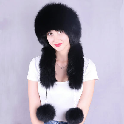 A woman wearing a large black fur ushanka hat with long ear flaps and two pom-poms, paired with a white t-shirt, against a plain gray studio background. The brand logo 'Joolsana' is visible in the top left corner.