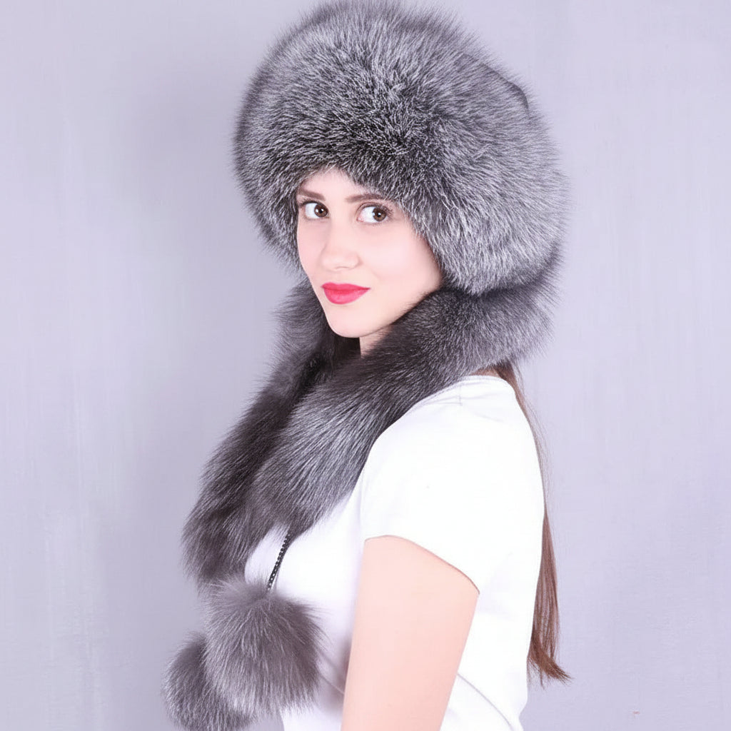 A woman wearing a large gray fur hat with matching fur trim and a matching fur neck warmer, photographed against a plain light gray background. The brand name 'Joelsana' appears in the upper left corner.