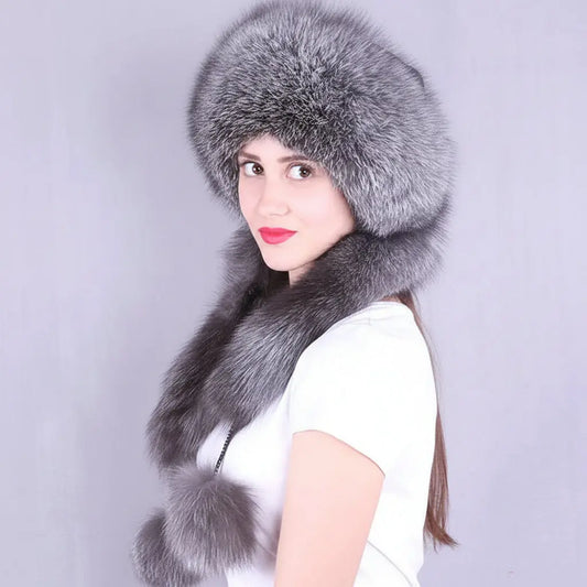 A woman wearing a large gray fur hat with matching fur trim and a matching fur neck warmer, photographed against a plain light gray background. The brand name 'Joelsana' appears in the upper left corner.