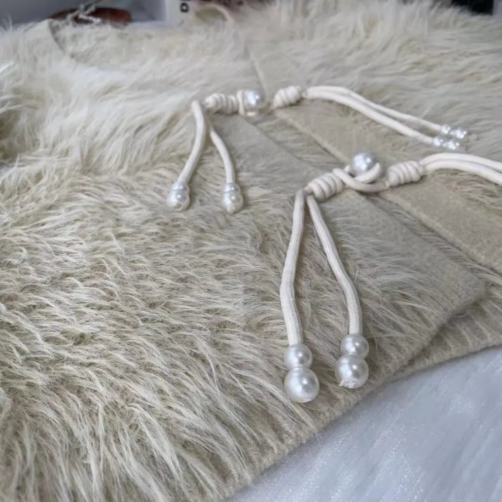 A close-up of a beige garment featuring cream-colored drawstrings adorned with faux pearls, laid over a fluffy cream faux fur surface.