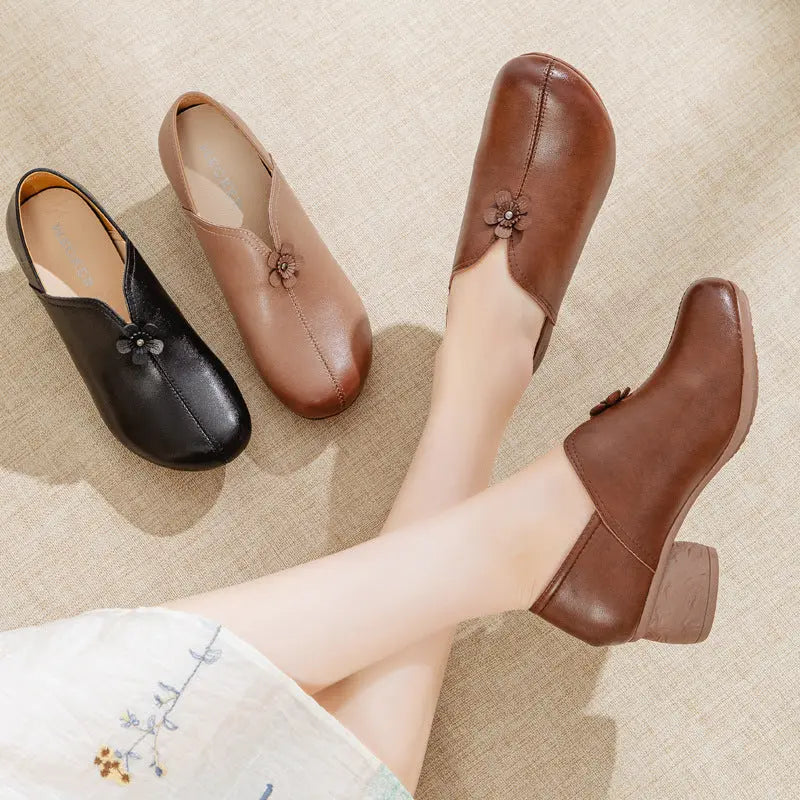 Four low-heeled slip-on shoes with floral embellishments on the front, shown on a textured beige floor. Three shoes are displayed as a flat lay, and one is being worn on a person's foot, with the other leg crossed over.