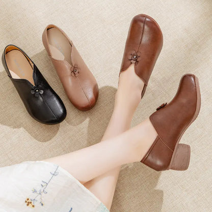Four low-heeled slip-on shoes with floral embellishments on the front, shown on a textured beige floor. Three shoes are displayed as a flat lay, and one is being worn on a person's foot, with the other leg crossed over.