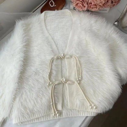 White faux fur cropped cardigan with cream cord and pearl button details, laid flat on a light surface with decorative flowers in the background.