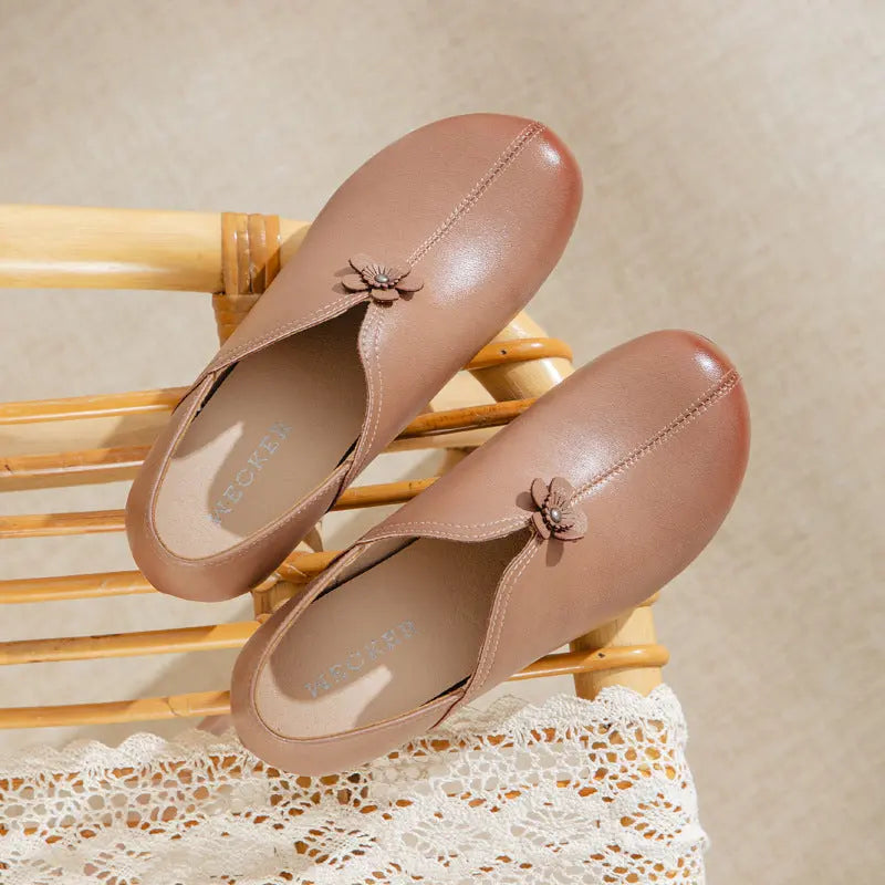 A pair of women's slip-on loafers in a warm taupe-brown color, styled on a light wood slatted chair over a white lace runner. Each shoe features a small decorative fabric flower accent on the vamp.
