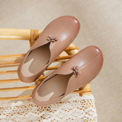 A pair of women's slip-on loafers in a warm taupe-brown color, styled on a light wood slatted chair over a white lace runner. Each shoe features a small decorative fabric flower accent on the vamp.