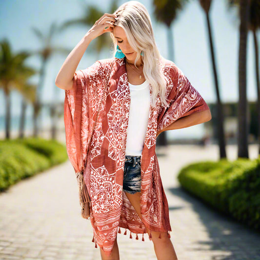 Woman wearing a patterned boho kimono-style cover-up in blue, paired with a wide-brimmed hat, white top, and denim shorts. Smaller images feature the same cover-up in black, teal, and rust colors—elegant, flowy, and perfect for layering.