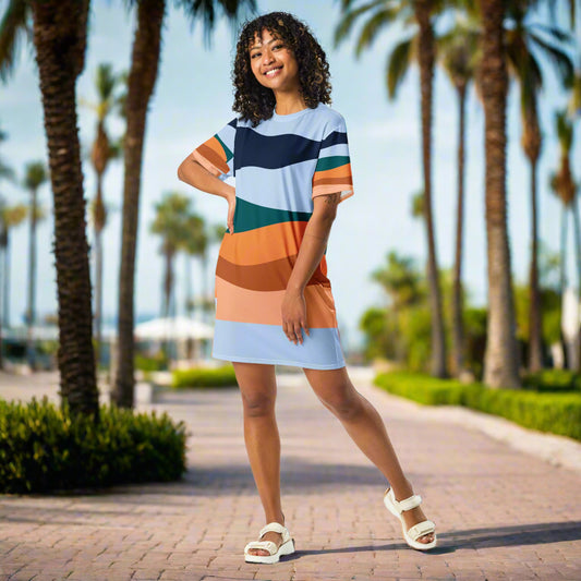 Person wearing a colorful striped t-shirt dress with shades of blue, orange, green, and brown, paired with white sandals, standing on a paved pathway lined with palm trees—stylish, vibrant, and perfect for sunny days.