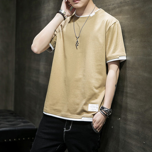 Person wearing a khaki t-shirt with white sleeve and hem accents, accessorized with a necklace, a watch, and bracelets, leaning casually against a dark textured wall—modern, stylish, and available in 8 colors.